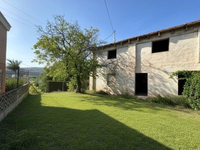 Vista exterior de la propiedad, mostrando su fachada de dos plantas con revestimiento claro y aberturas sin carpintería. El amplio jardín con césped y un árbol maduro ofrece un lienzo para el diseño.