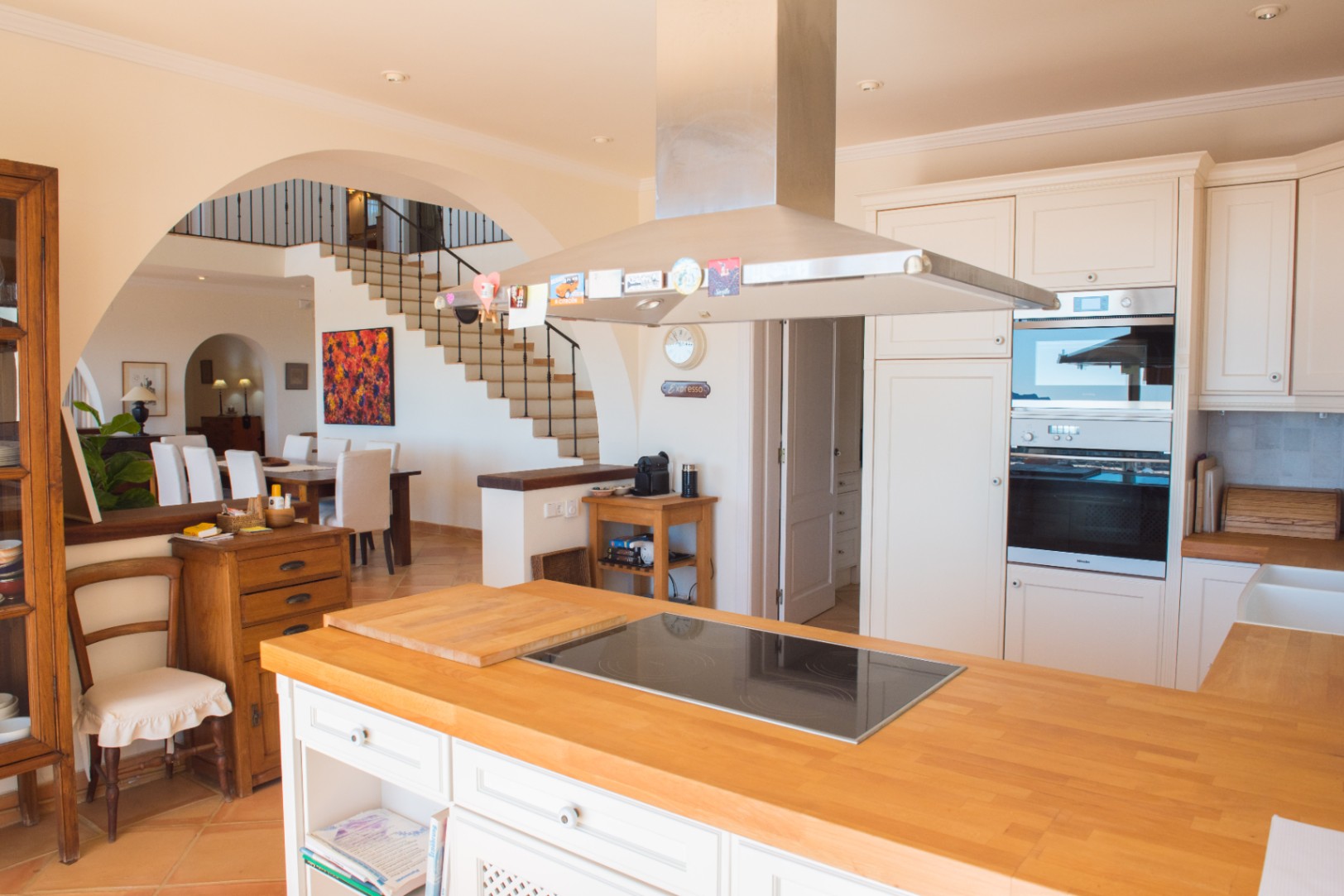 Cocina de concepto abierto con isla de madera, placa de inducción, campana de acero inoxidable y hornos integrados, conectada al comedor mediante un arco.