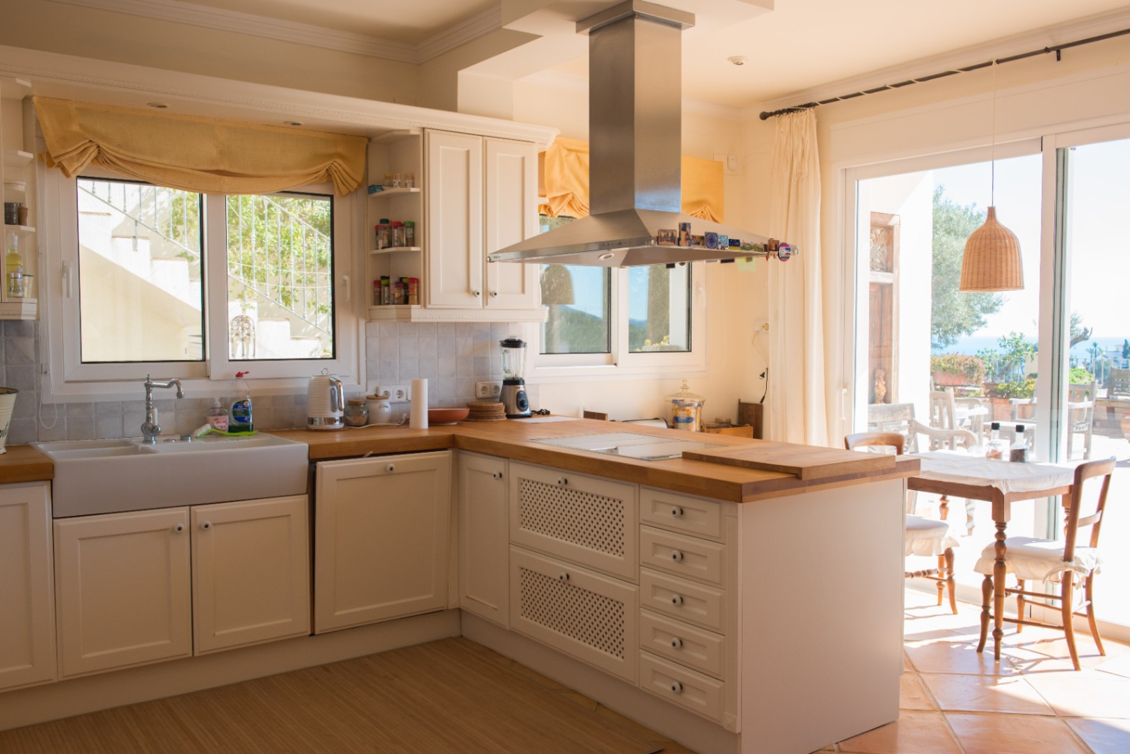 Cocina de estilo rústico con armarios blancos, encimeras de madera maciza, fregadero cerámico y campana de acero inoxidable con acceso directo a la terraza.