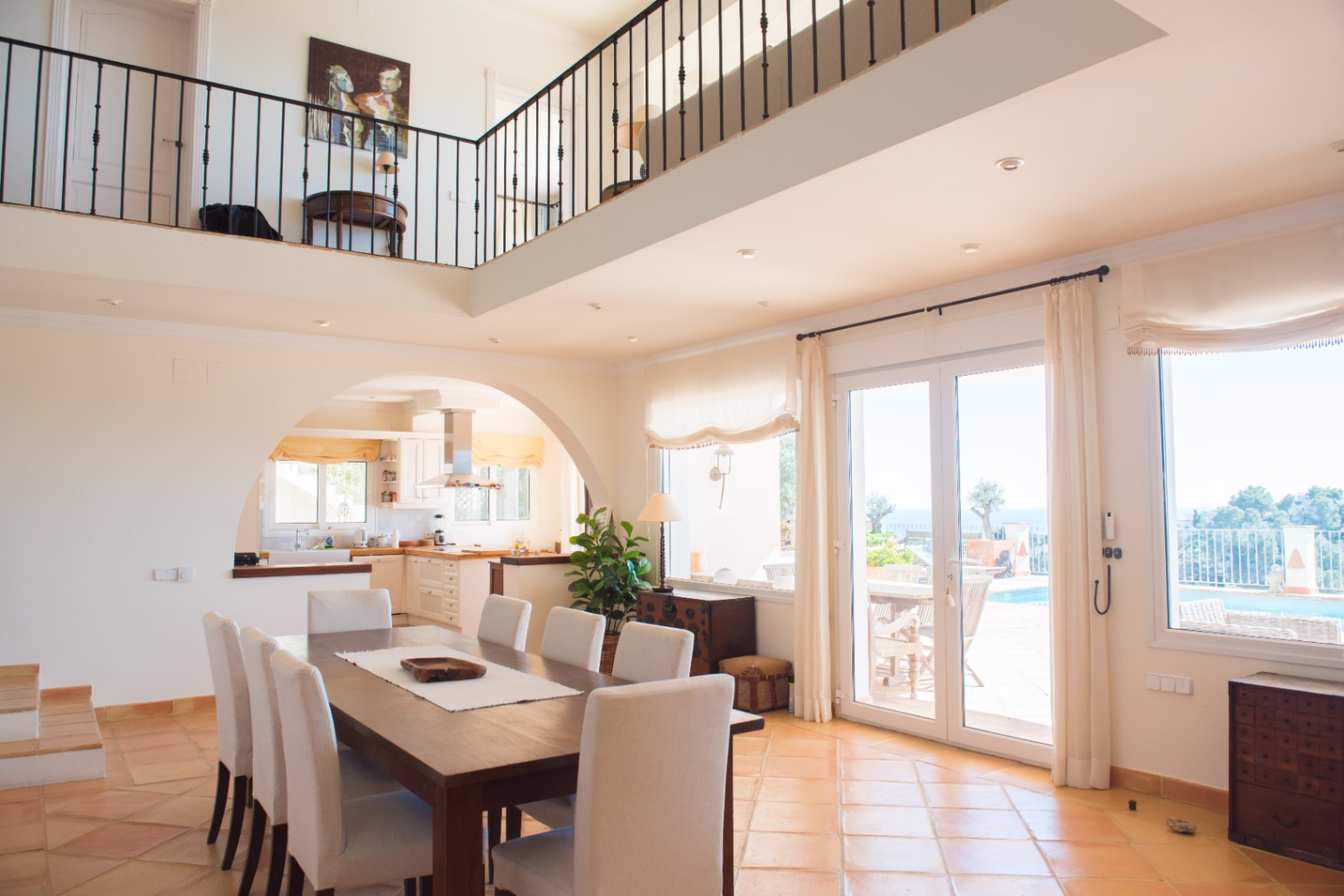 Comedor amplio con suelos de terracota, mesa de madera y acceso en arco a la cocina. Una galería superior con barandilla de hierro forjado domina la estancia.