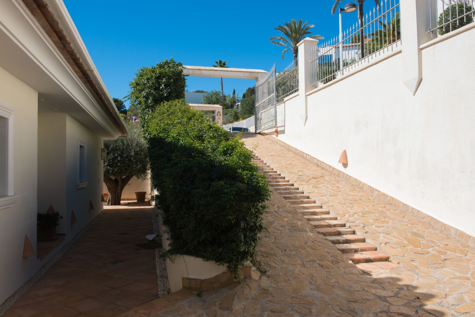 Entrada exterior con rampa de piedra natural y escalones integrados, flanqueada por muros blancos decorados con apliques y setos mediterráneos.