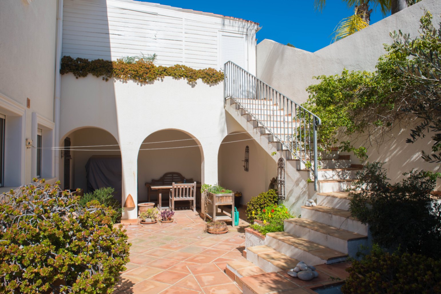 Patio exterior con pavimento de barro cocido, porches con arcos de medio punto y una escalera de obra con barandilla metálica hacia la planta superior.