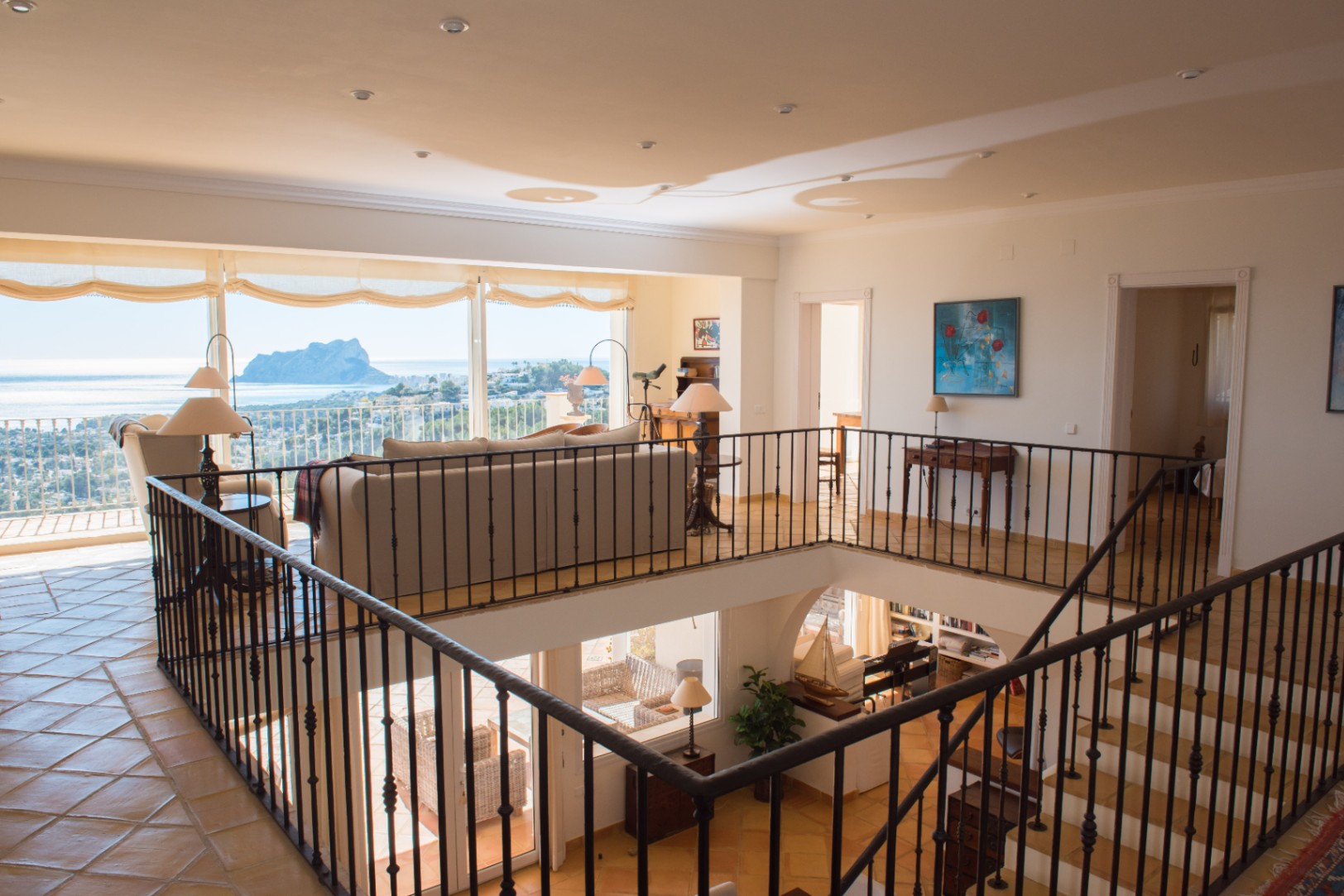 Salón en planta superior con barandilla de hierro forjado, suelos cerámicos y amplios ventanales con vistas panorámicas al Peñón de Ifach y el mar.