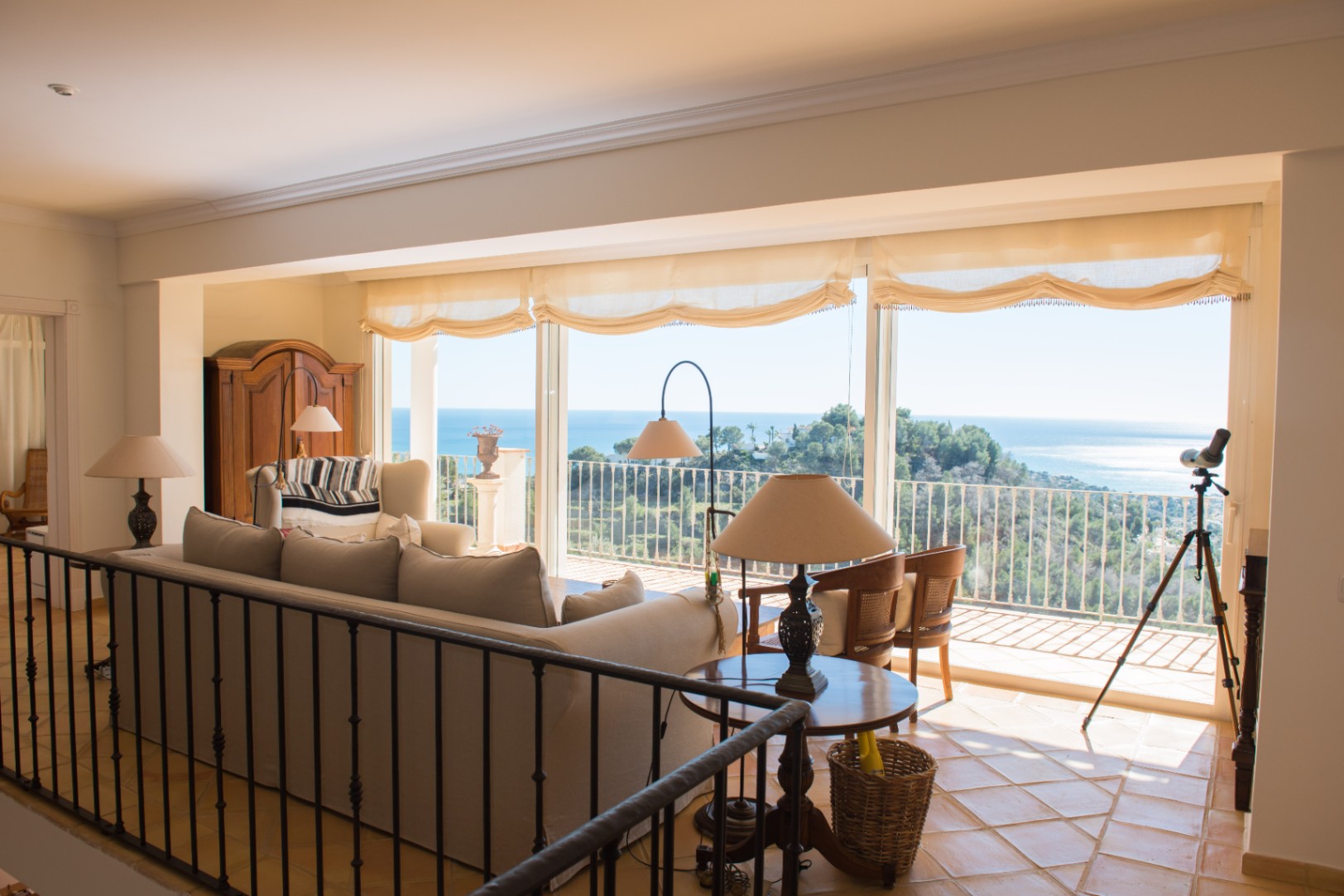 Salón luminoso con suelos de terracota, barandilla de hierro forjado y amplios ventanales que ofrecen vistas panorámicas al mar Mediterráneo y la costa.