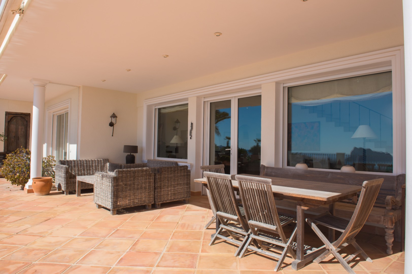 Terraza cubierta con suelos de terracota, zona de comedor de madera y mobiliario de mimbre, conectada al interior mediante amplios ventanales.