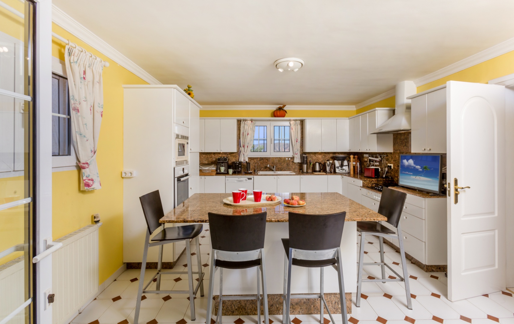 Cocina espaciosa con isla central de granito, taburetes negros, armarios blancos y paredes amarillas. El suelo cerámico presenta detalles decorativos en tono marrón.