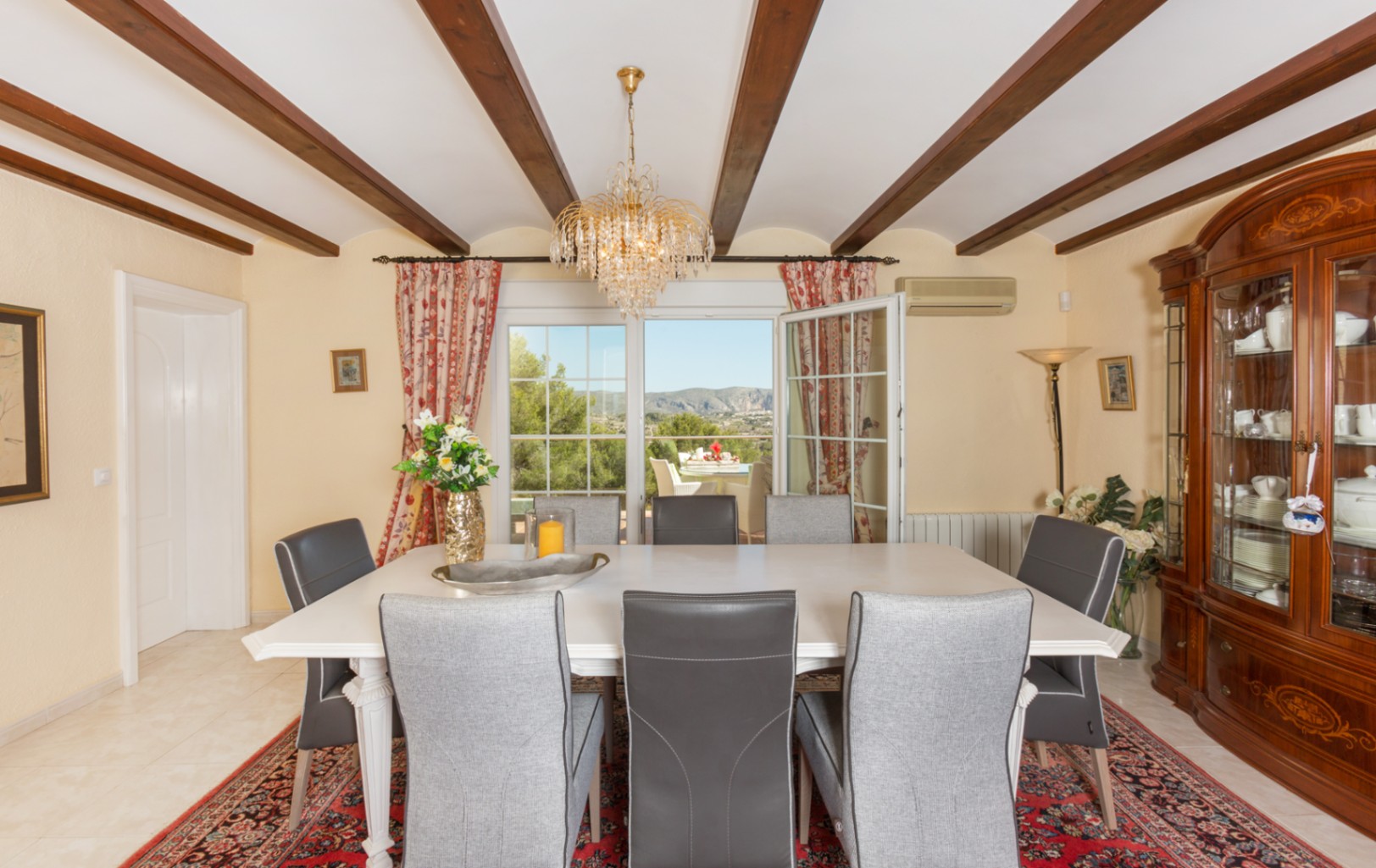 Comedor con vigas de madera vistas, mesa blanca y sillas tapizadas. Incluye una vitrina clásica y acceso a la terraza con vistas a las montañas.