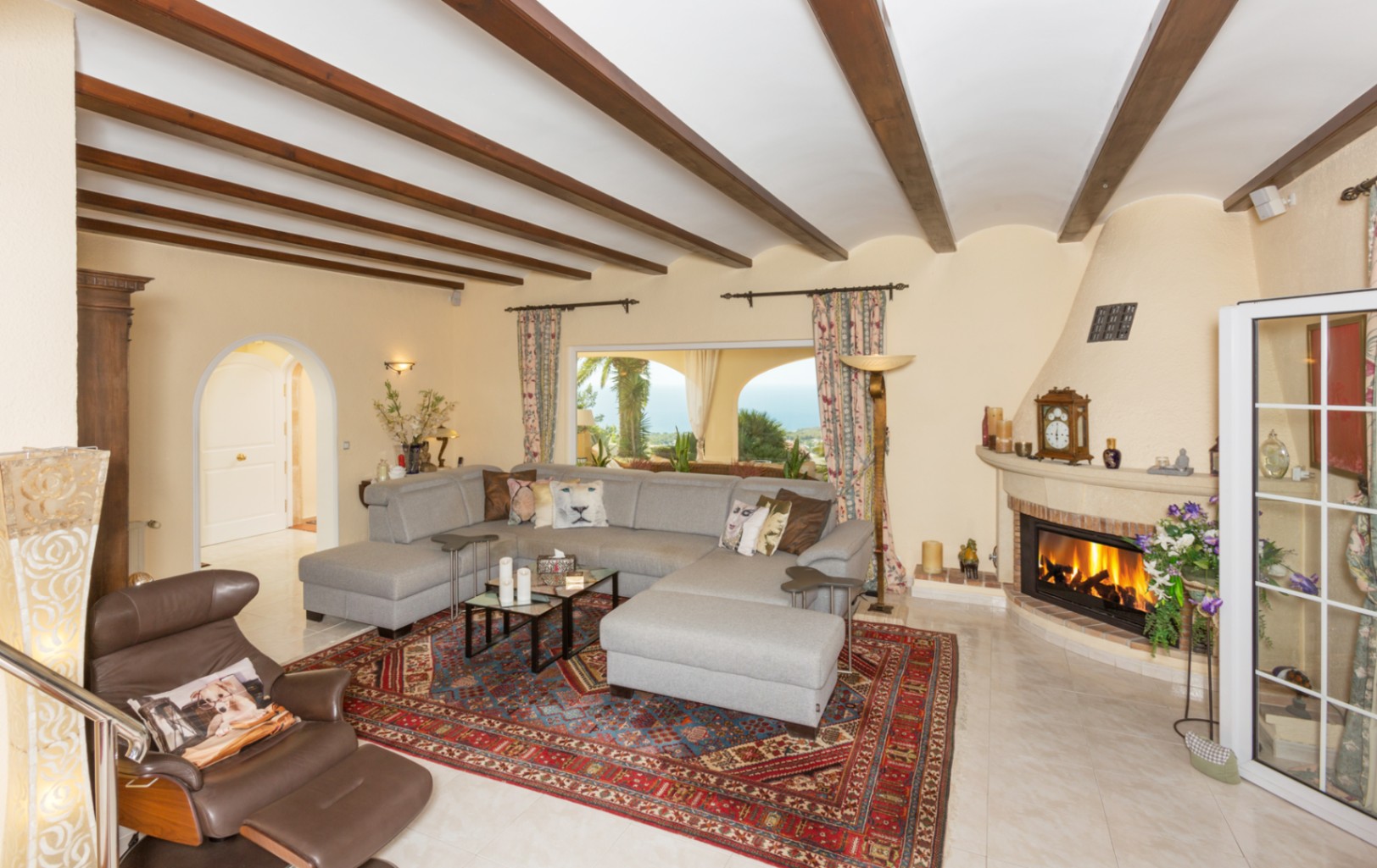 Salón espacioso con vigas de madera a la vista, gran sofá rinconero gris, chimenea de obra y vistas al mar a través del ventanal principal.