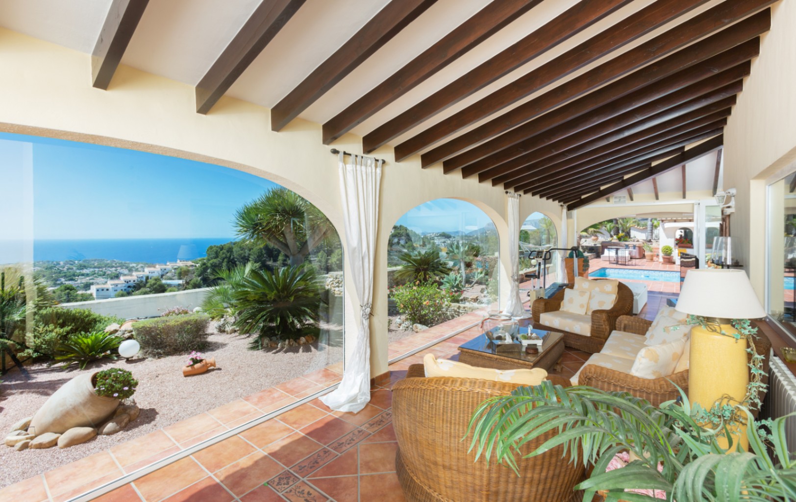 Terraza cubierta con vigas de madera oscura, suelos de terracota y mobiliario de mimbre. Los arcos acristalados ofrecen vistas panorámicas al mar y al jardín mediterráneo.