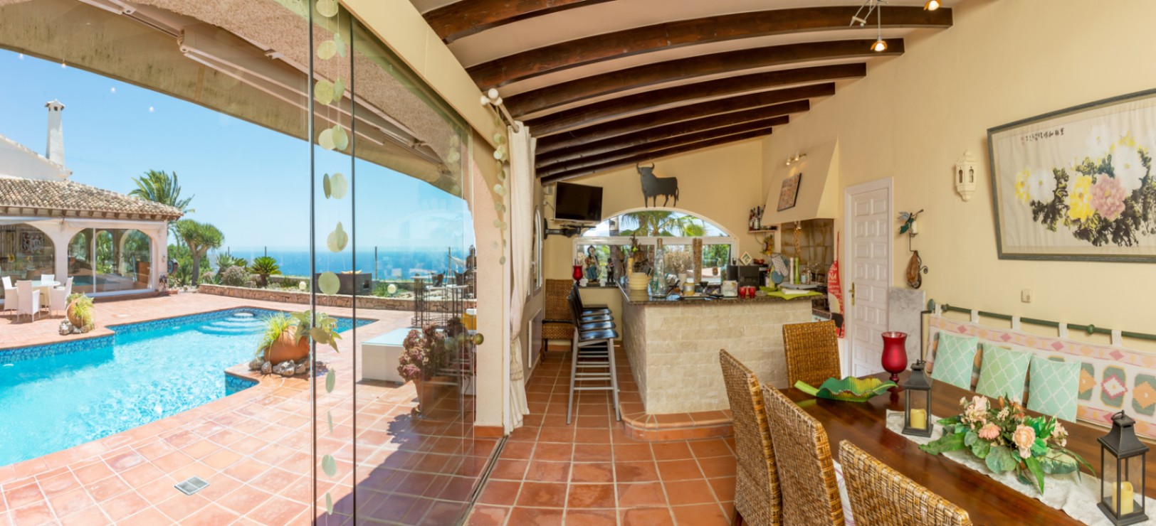 Terraza cubierta con vigas de madera, suelo de terracota y cocina exterior con barra de piedra, conectada a la piscina mediante grandes ventanales de cristal.