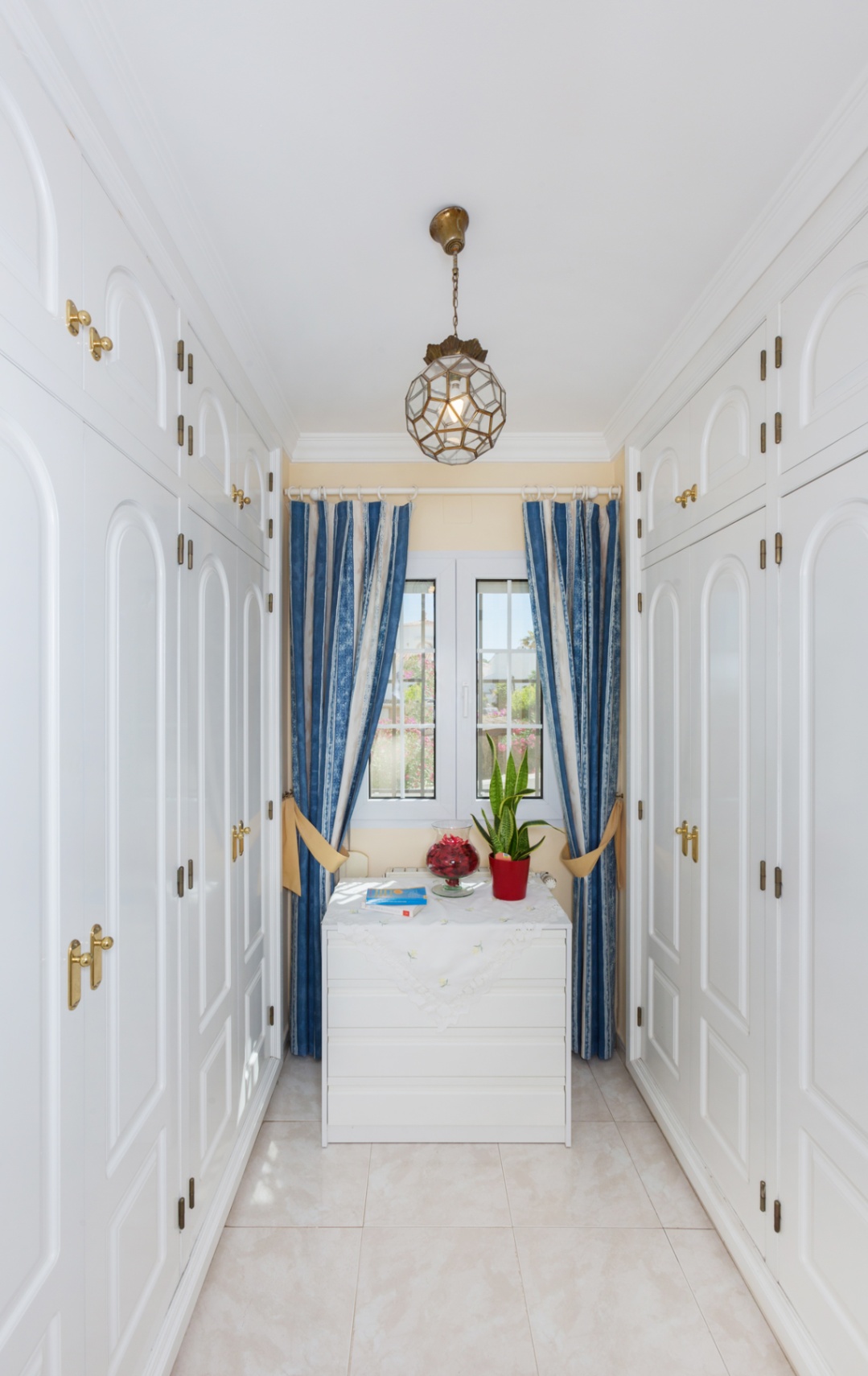 Vestidor con armarios empotrados blancos de suelo a techo, herrajes de latón, ventana central con cortinas a rayas azules y una lámpara colgante geométrica.