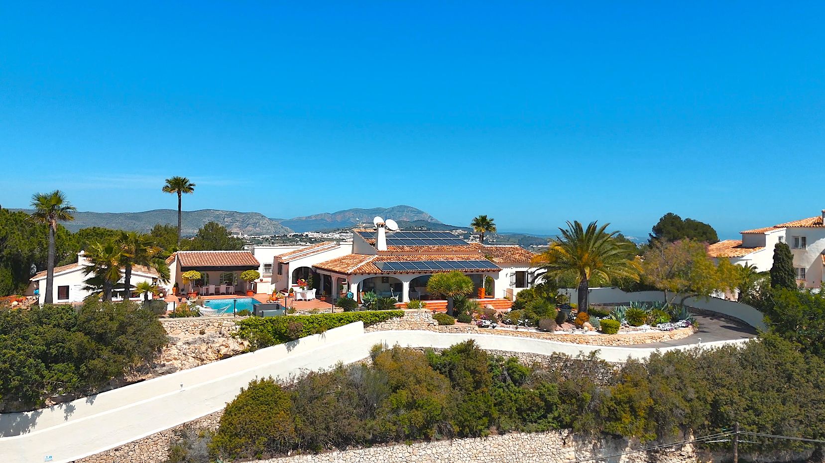Vista aérea de la villa con tejados de terracota, paneles solares y piscina, rodeada de jardines mediterráneos con vistas panorámicas al mar.
