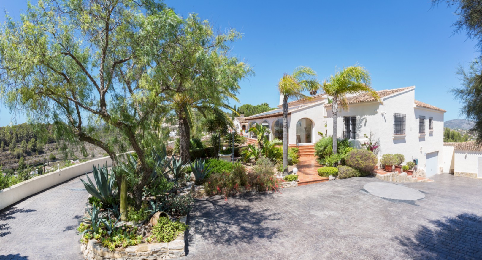 Vista exterior de la villa mediterránea con entrada pavimentada, jardines con agaves y palmeras, y fachada blanca con detalles de arcos y rejas de hierro.