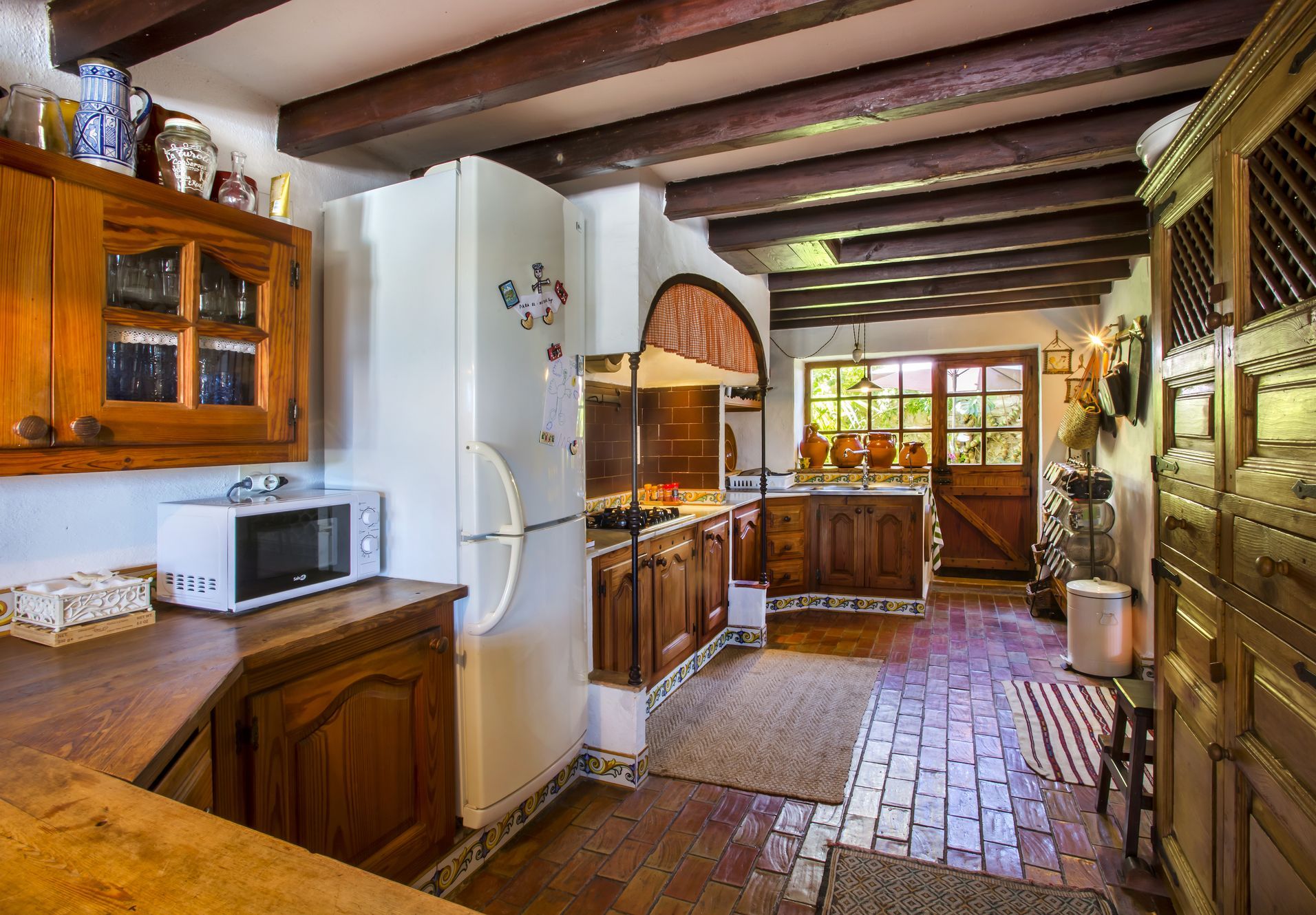 Cocina rústica con suelos de terracota, vigas de madera y muebles de madera oscura. La luz natural entra por la puerta acristalada.