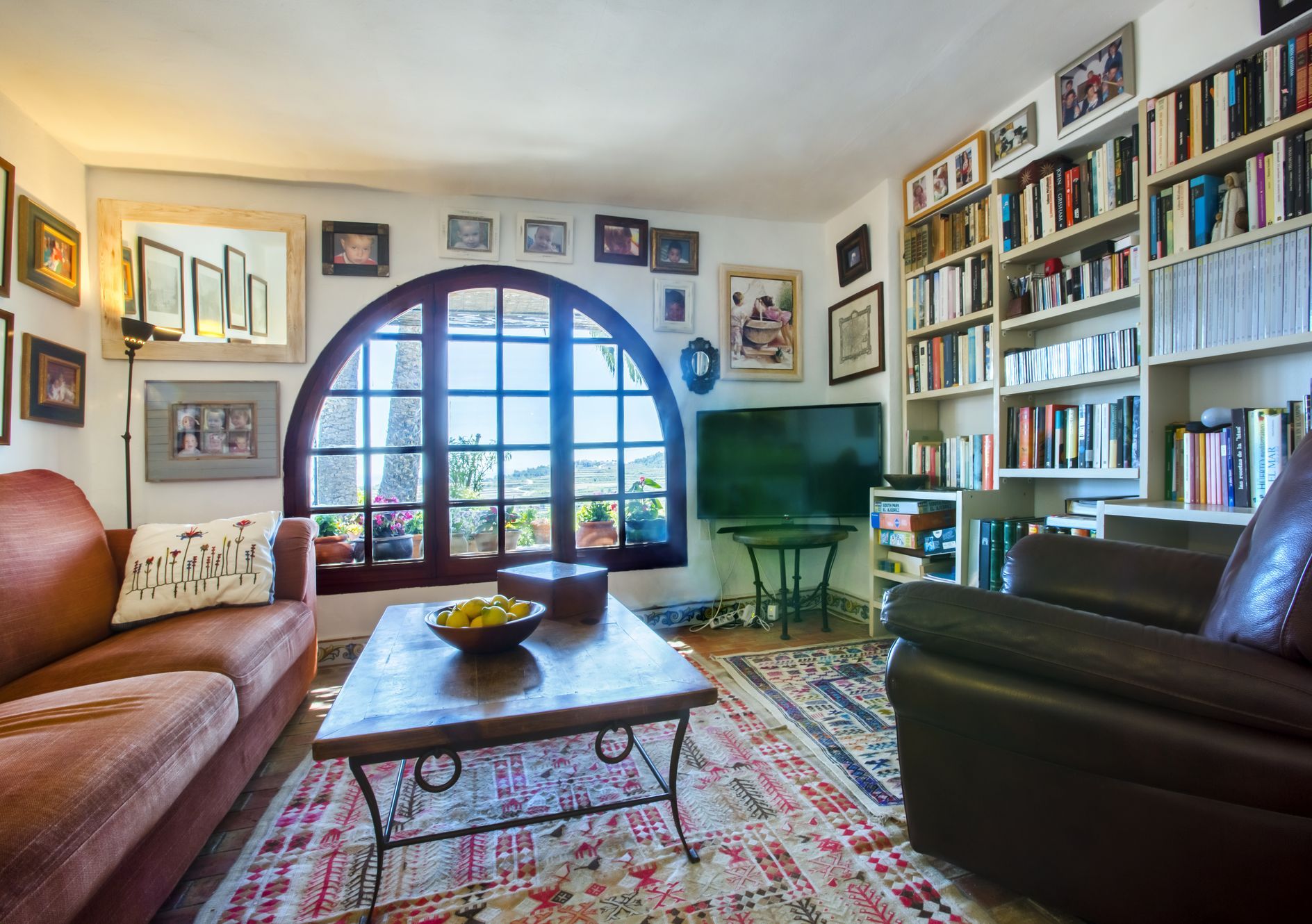 Salón con ventana arqueada y vistas, decorado con alfombra estampada y estantería llena de libros.