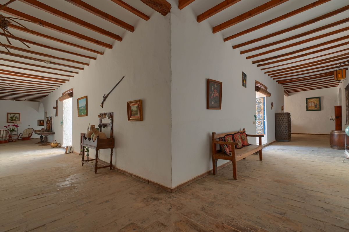 Amplio distribuidor con vigas de madera vistas, suelos de piedra rústica y paredes blancas decoradas con mobiliario antiguo y elementos históricos tradicionales.