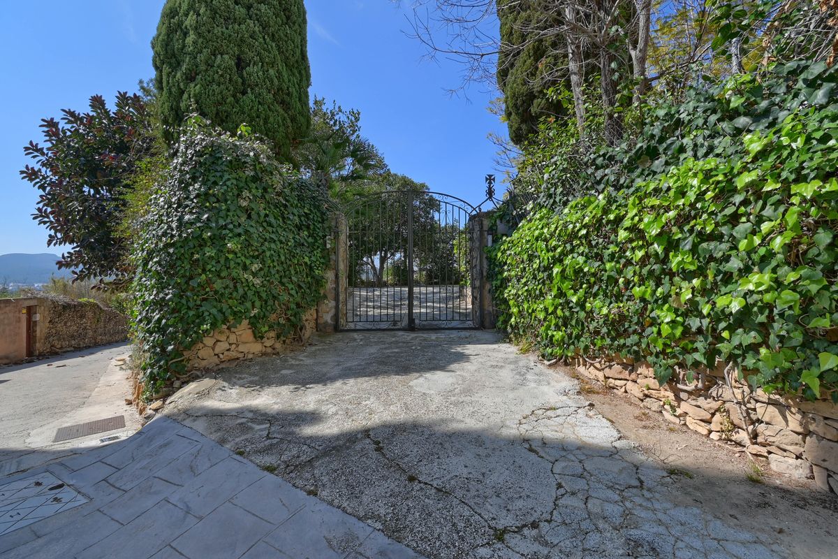 Entrada principal con portón de hierro forjado negro, muros de piedra natural cubiertos de hiedra y cipreses maduros que garantizan privacidad.