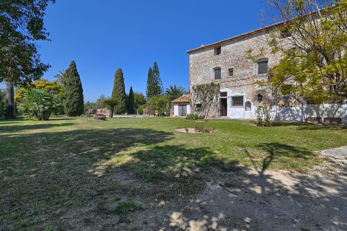 Fachada de piedra natural de la finca histórica rodeada de extensos jardines con césped, cipreses y palmeras bajo un cielo despejado.