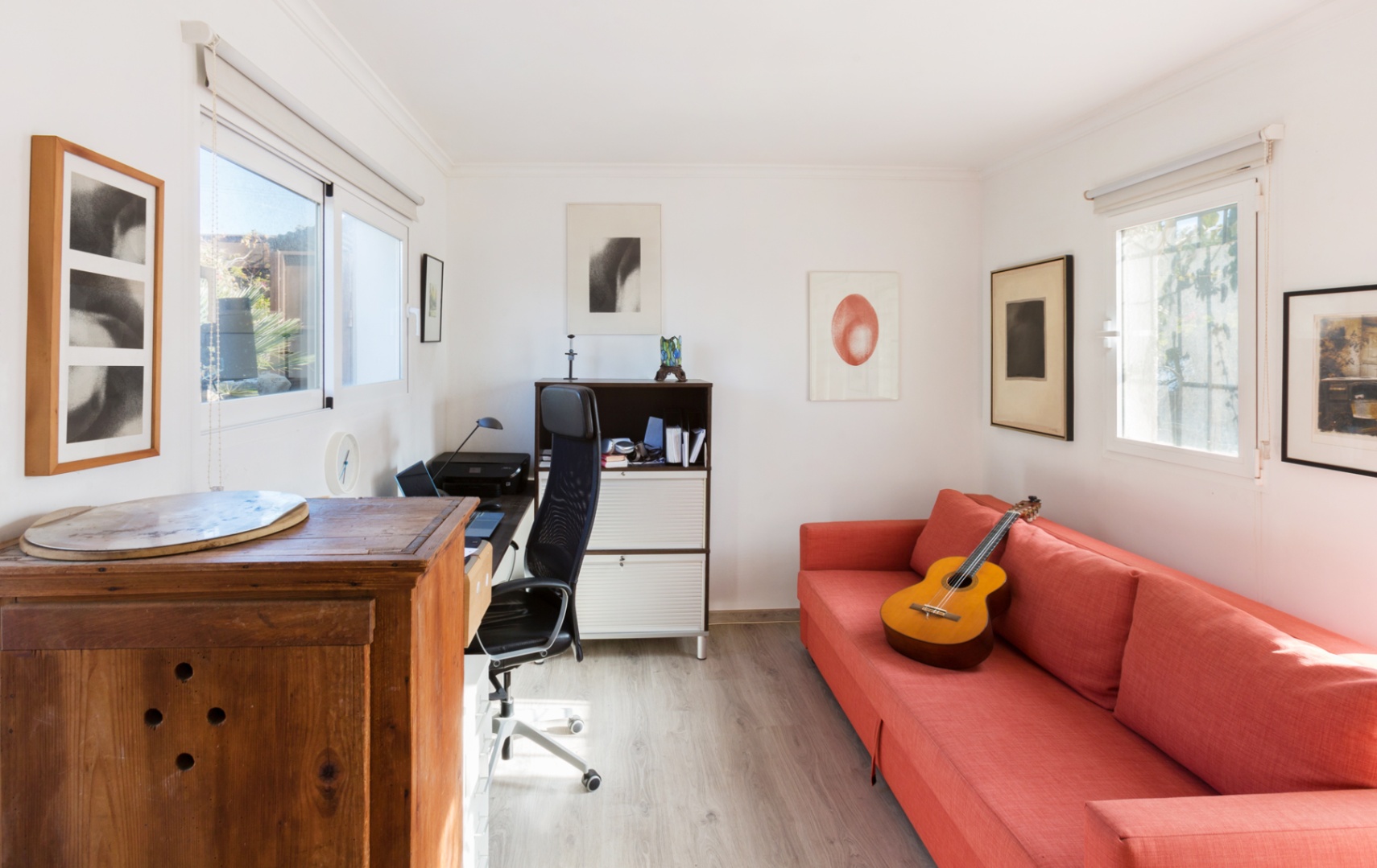 Despacho luminoso con suelos de madera clara, sofá rojo, zona de trabajo con silla ergonómica y luz natural a través de ventanas con estores.