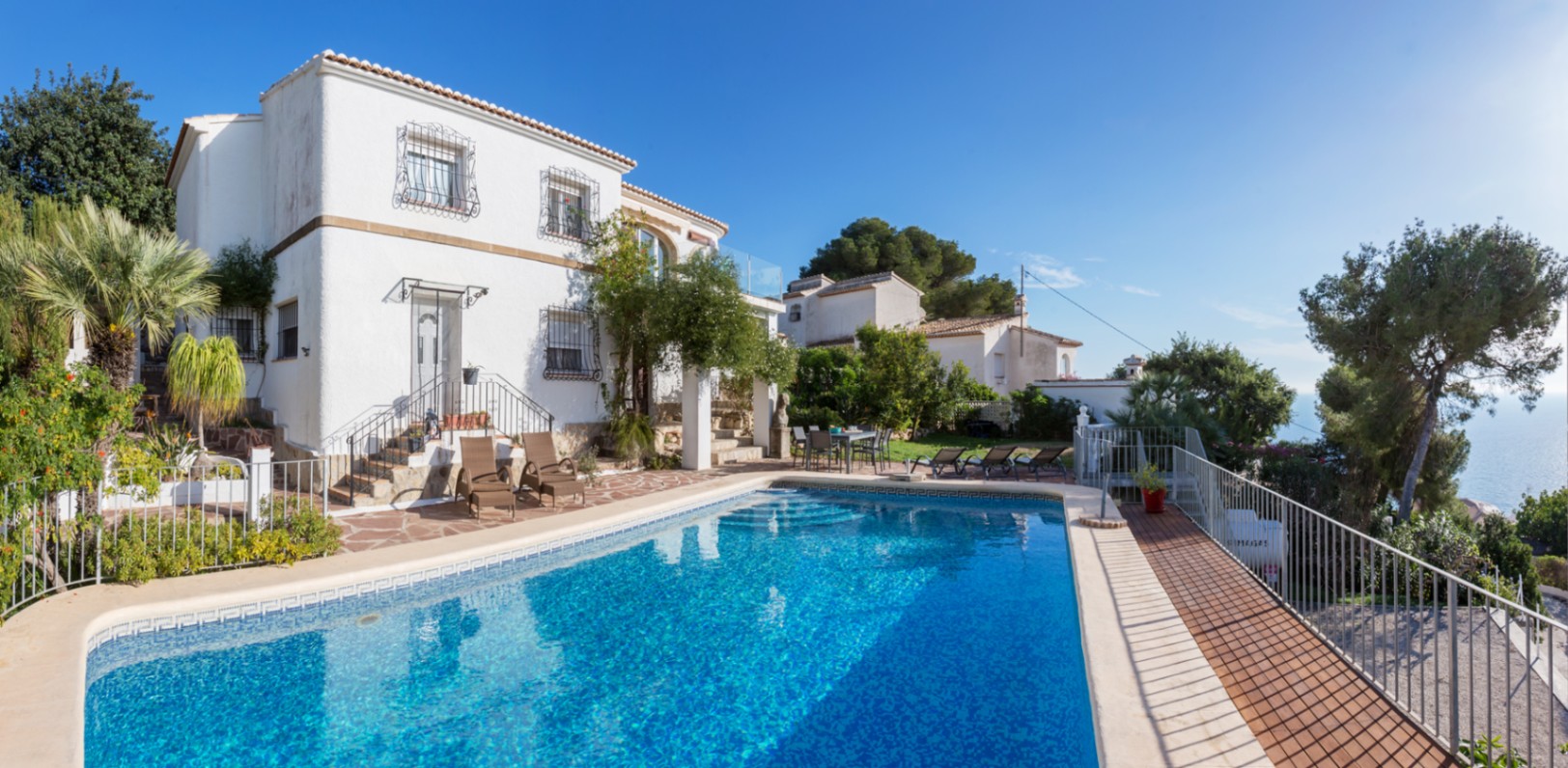 Exterior de la villa mediterránea con piscina rectangular, terraza de piedra con tumbonas y fachada blanca con rejas tradicionales frente a las vistas al mar.