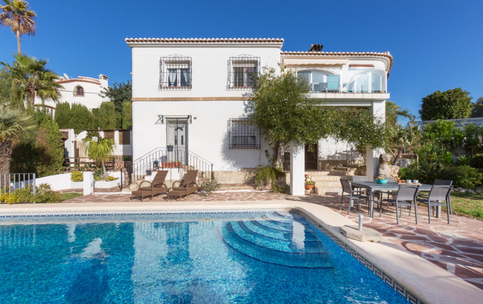 Fachada exterior de la villa con piscina privada, terraza de piedra natural con zona de comedor y ventanas protegidas por rejas de hierro forjado.