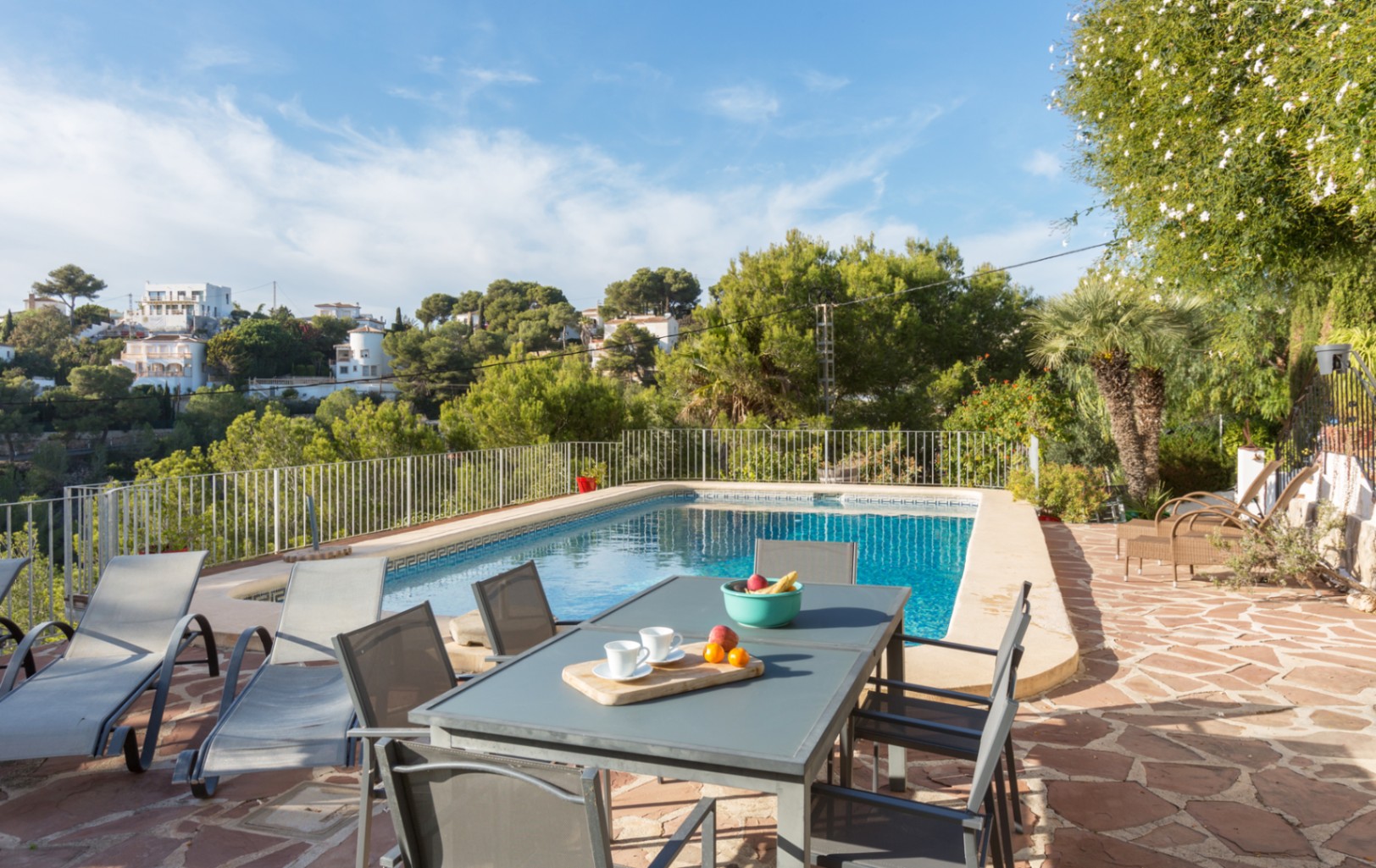 Terraza exterior con pavimento de piedra natural, piscina rectangular y mobiliario de comedor, delimitada por una barandilla metálica y pinos mediterráneos.