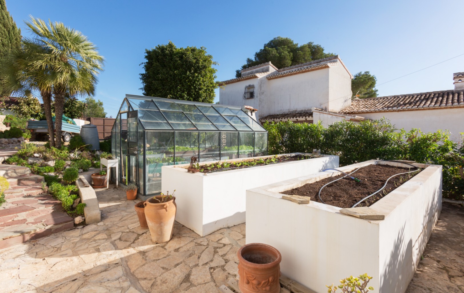 Zona de jardín con invernadero de cristal y dos grandes jardineras elevadas blancas con riego integrado sobre un pavimento de piedra natural irregular.