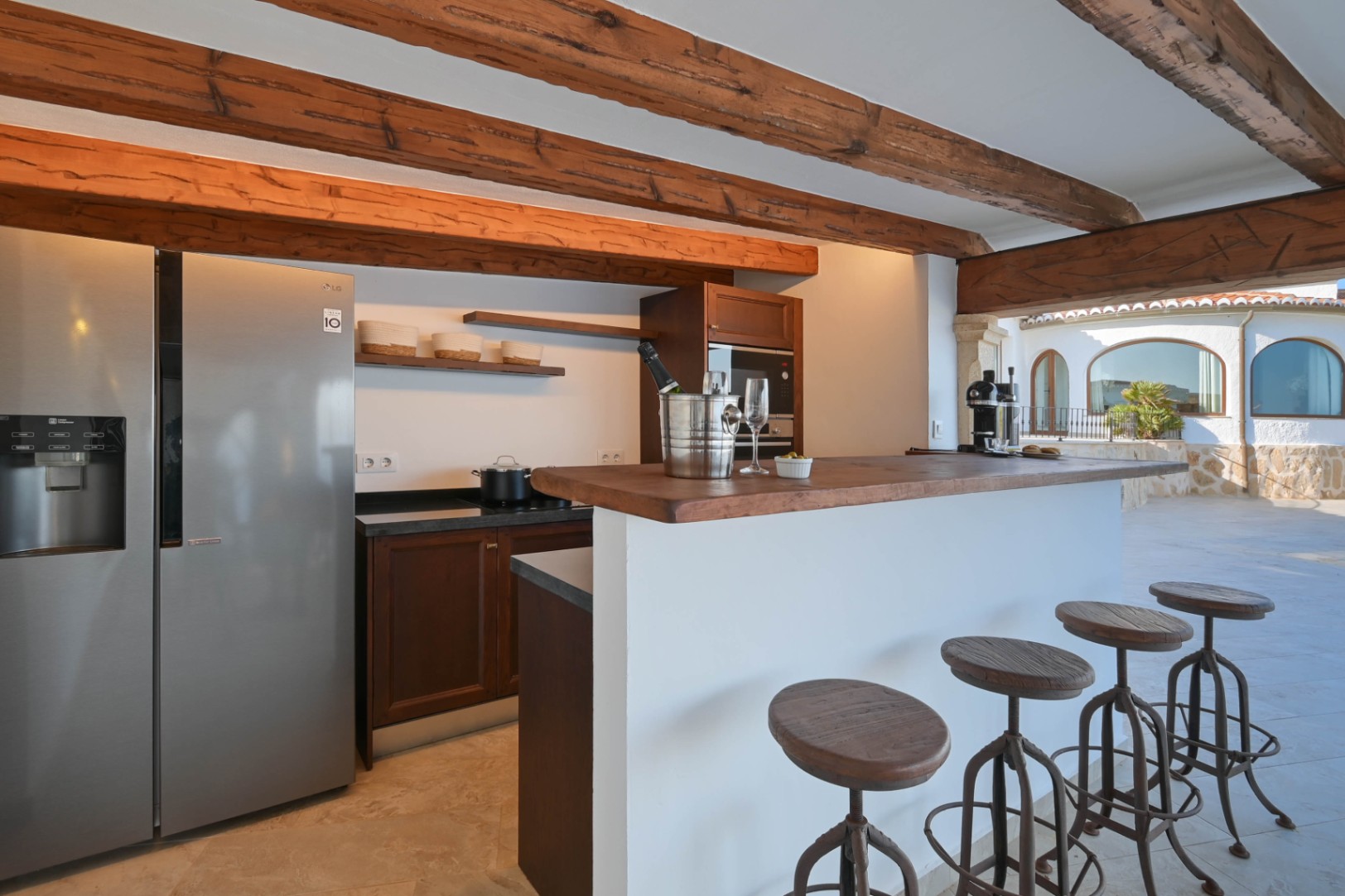 Cocina rústica con vigas de madera vista, frigorífico de acero inoxidable y barra americana blanca. Incluye taburetes industriales y vistas al patio exterior con muros de piedra.