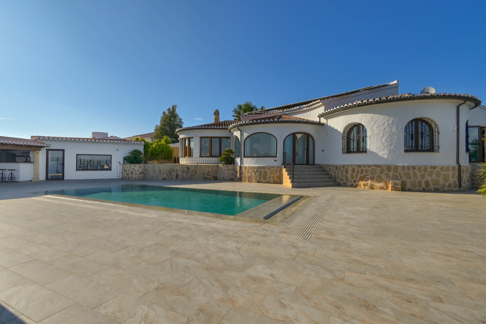 Exterior de una villa mediterránea con piscina rectangular, zócalo de piedra natural y amplia terraza pavimentada bajo un cielo despejado.