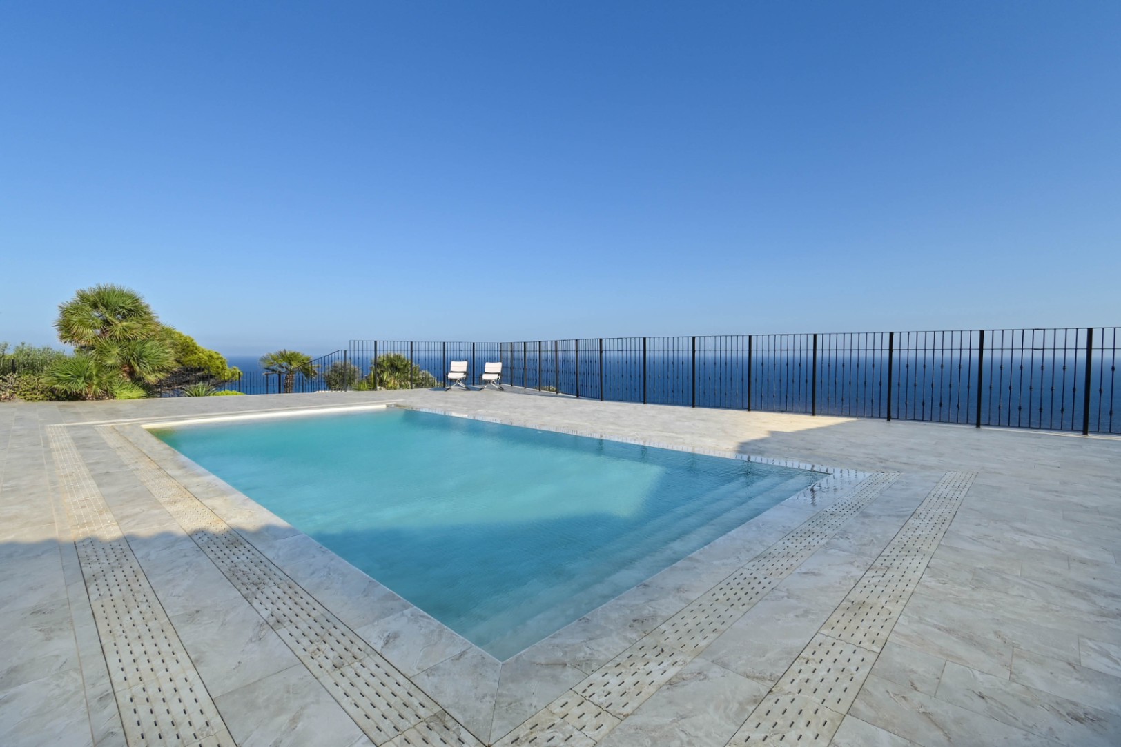 Piscina rectangular con escalones integrados, situada en una terraza de piedra clara con barandilla metálica y vistas panorámicas al mar.