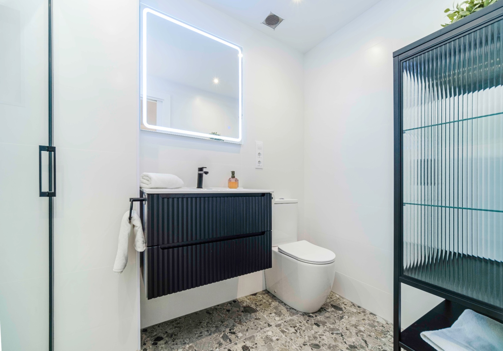 Baño moderno con mueble suspendido negro estriado, grifería mate y espejo retroiluminado sobre suelo de terrazo gris.