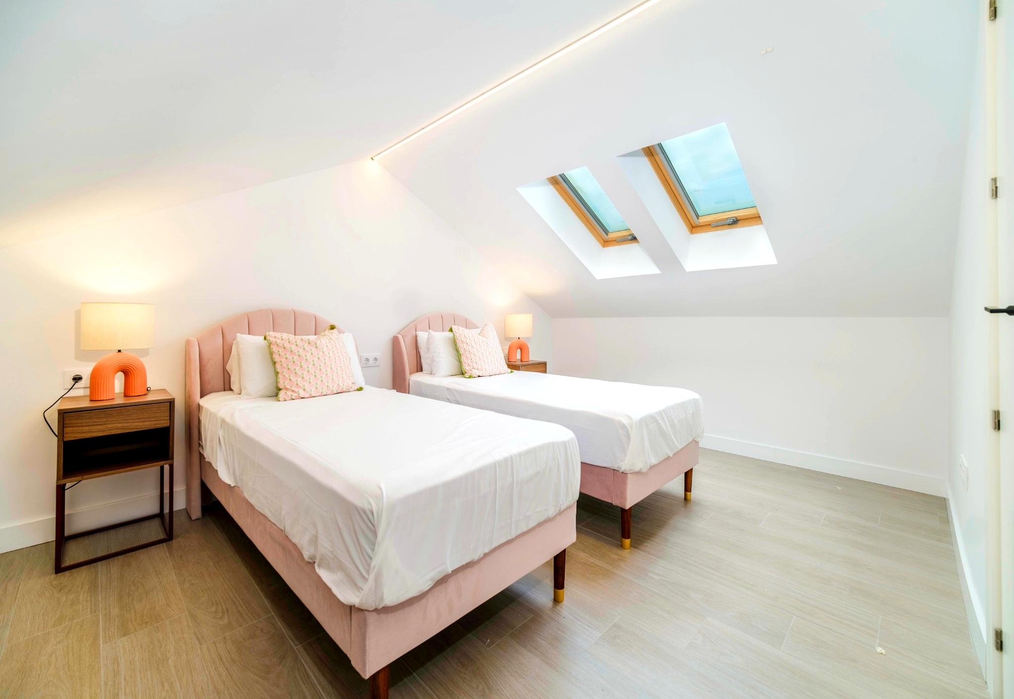Dormitorio abuhardillado con dos camas individuales de terciopelo rosa, claraboyas dobles y una tira de iluminación LED integrada en el techo inclinado.