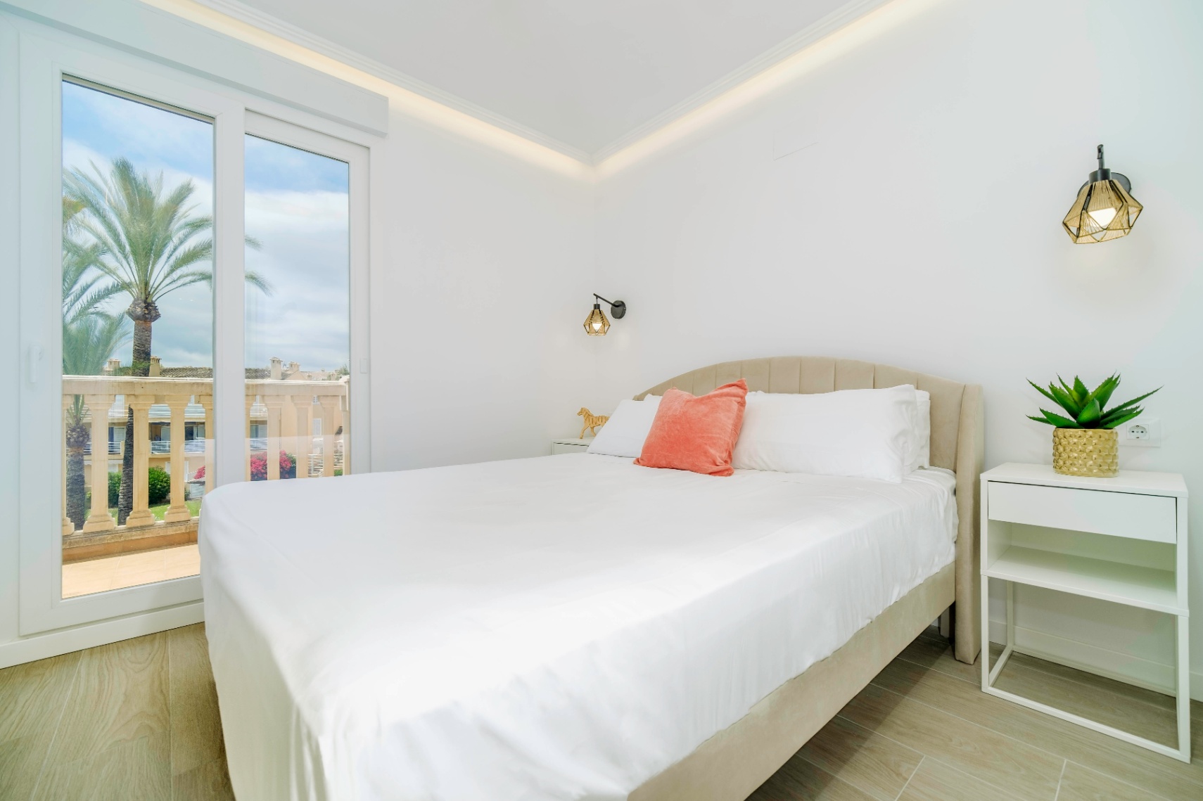 Dormitorio moderno con cama de cabecero tapizado, apliques geométricos y acceso directo a una terraza con vistas a palmeras y balaustrada de piedra.