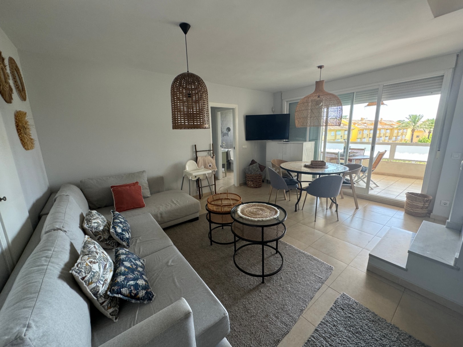 Salón comedor luminoso con sofá rinconero gris, lámparas de fibras naturales y mesa de comedor circular. Acceso directo a una terraza amueblada a través de ventanales.