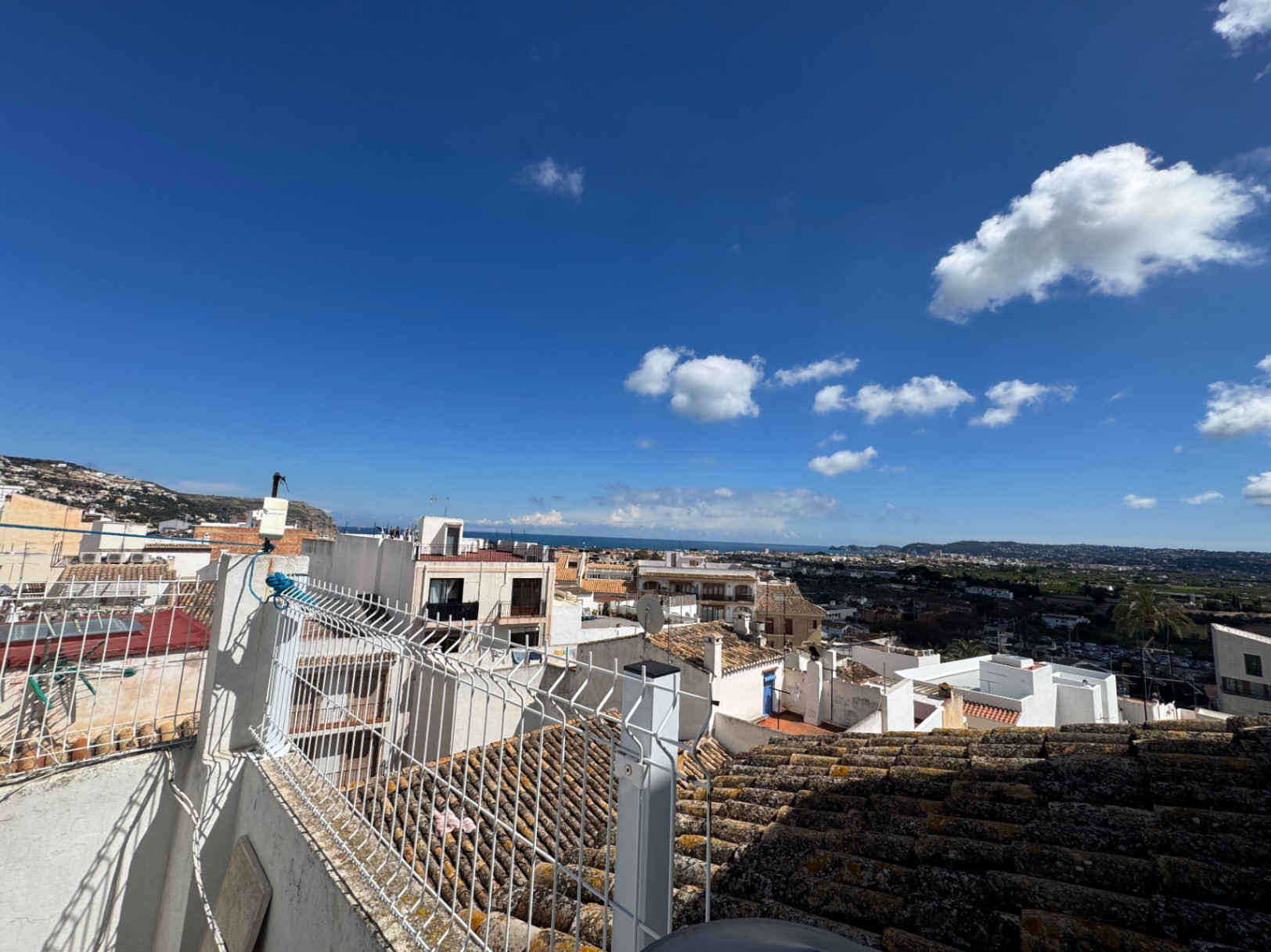 Vista elevada desde una terraza sobre edificios tradicionales de paredes blancas y tejados de terracota, con el mar Mediterráneo visible en el horizonte bajo un cielo despejado.