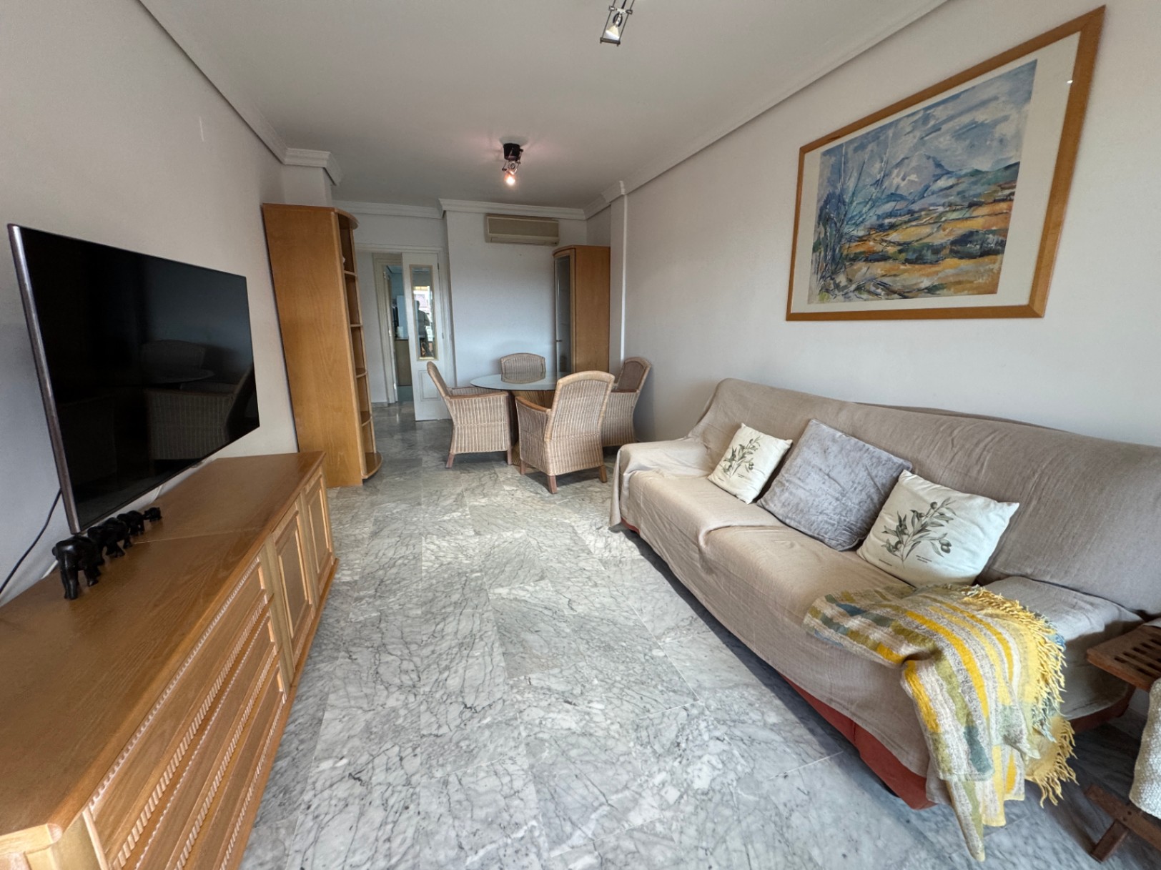 Salón comedor con suelos de mármol gris, mueble de televisión en madera y zona de comedor al fondo con mesa de cristal y sillas de mimbre.
