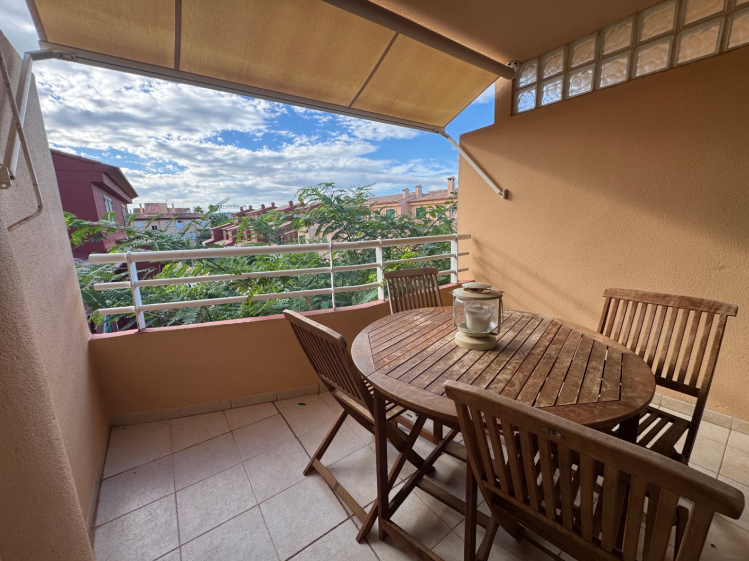 Terraza cubierta con conjunto de comedor de madera, toldo extensible y suelo cerámico, con vistas a zonas verdes y edificios residenciales circundantes.