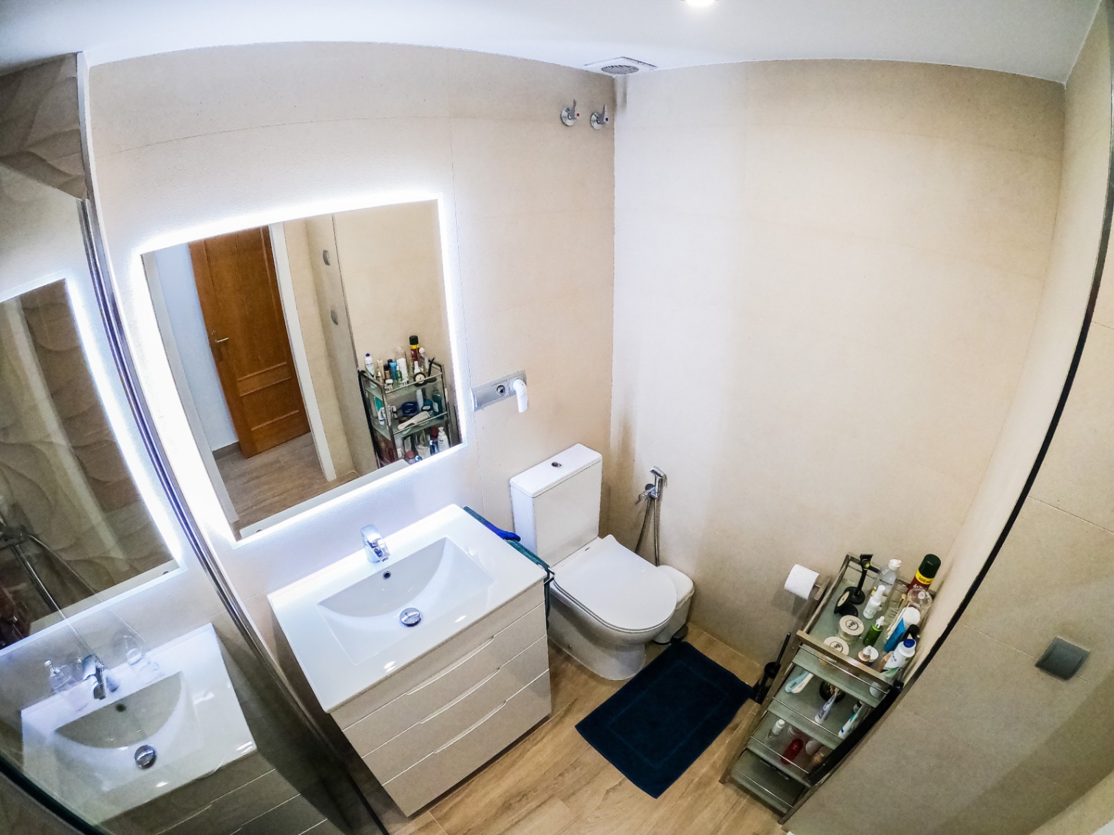 Baño moderno con mueble de lavabo blanco, espejo retroiluminado por LED y suelo cerámico imitación madera. Incluye inodoro y paredes revestidas en tonos beige neutros.