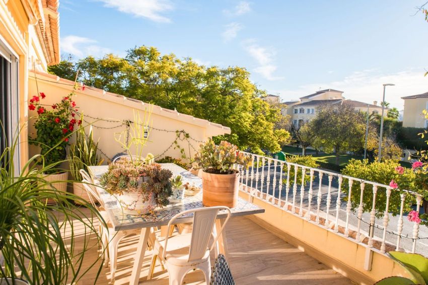 Soleada terraza con mesa de mosaico, sillas de metal blanco y diversas plantas en macetas. Incluye luces decorativas y vistas a la zona residencial.