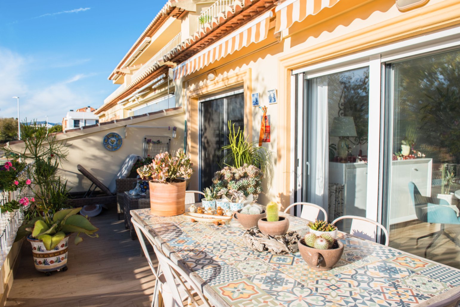 Terraza mediterránea soleada con mesa de comedor de mosaico, suculentas en macetas y amplios ventanales correderos bajo un toldo de rayas.