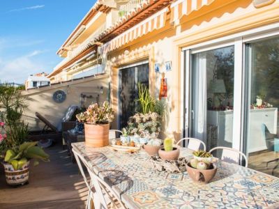 Amplia terraza soleada con mesa de comedor de azulejos de mosaico, diversas plantas suculentas en macetas y cómodos asientos. Toldos a rayas ofrecen sombra y acceso directo al interior.