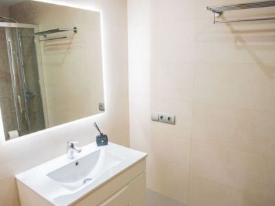 Baño moderno con mueble blanco y lavabo integrado, espejo iluminado y paredes de azulejos beige. Se observa una ducha acristalada en el reflejo.