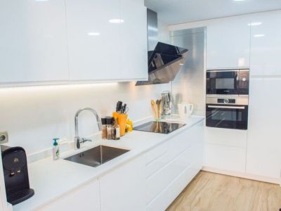 Cocina moderna con armarios blancos de alto brillo, electrodomésticos Bosch integrados y encimera blanca. Iluminación LED bajo los armarios destaca el fregadero de acero y la placa de inducción.