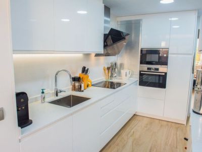 Cocina moderna con armarios blancos de alto brillo, encimera clara y electrodomésticos Bosch integrados. Incluye placa de inducción, fregadero de acero inoxidable e iluminación bajo mueble.