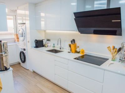 Cocina moderna con armarios blancos de alto brillo, placa de inducción negra y campana extractora angular. Incluye iluminación bajo mueble y lavadora/secadora integradas junto a la ventana.