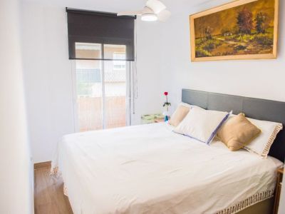 Dormitorio con cama doble y cabecero tapizado gris, suelo de madera y amplio ventanal con estor oscuro. Un ventilador de techo y un cuadro paisajístico decoran la estancia.