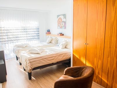 Dormitorio luminoso con dos camas individuales, armario de madera natural y suelos de tonos claros. La luz natural entra por las persianas verticales, iluminando el espacio.