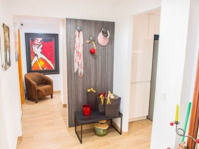 La entrada presenta un suelo de imitación madera clara y un moderno panel oscuro con ganchos. Un nicho adyacente muestra un sillón marrón y arte vibrante, con la cocina visible a la derecha.
