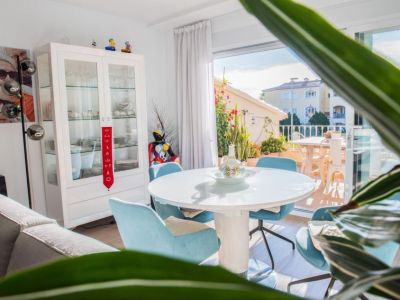 Luminoso comedor con mesa redonda blanca moderna y sillas azul claro. Grandes puertas correderas de cristal conectan con una terraza llena de plantas, aportando luz natural y vistas residenciales.