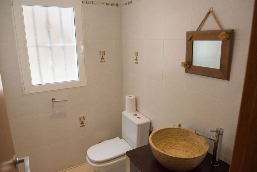 Aseo de cortesía con un distintivo lavabo de piedra natural sobre mueble de madera oscura y grifo moderno. Paredes alicatadas en blanco y espejo rústico de madera con cuerda. Aseo de cortesía con un distintivo lavabo de piedra natural sobre mueble de madera oscura y grifo moderno. Paredes alicatadas en blanco y espejo rústico de madera con cuerda.